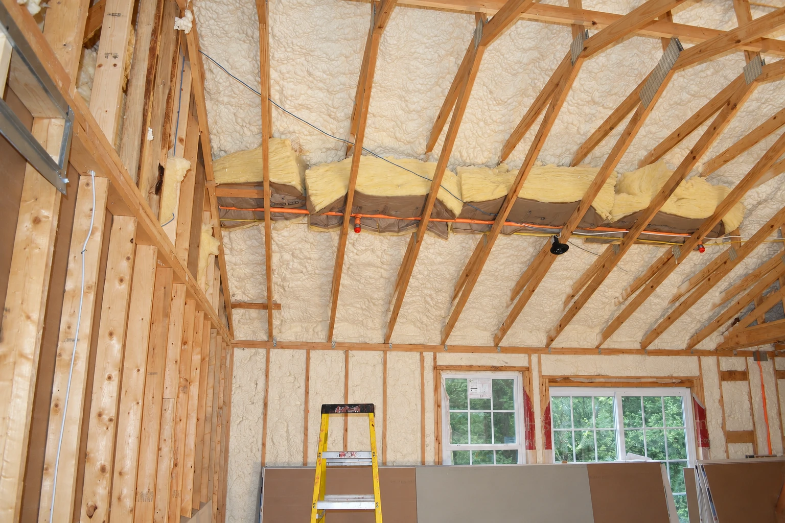 Unfinished attic with spray foam covering the roof and walls, exposed wooden trusses, and fiberglass batts resting across a beam.