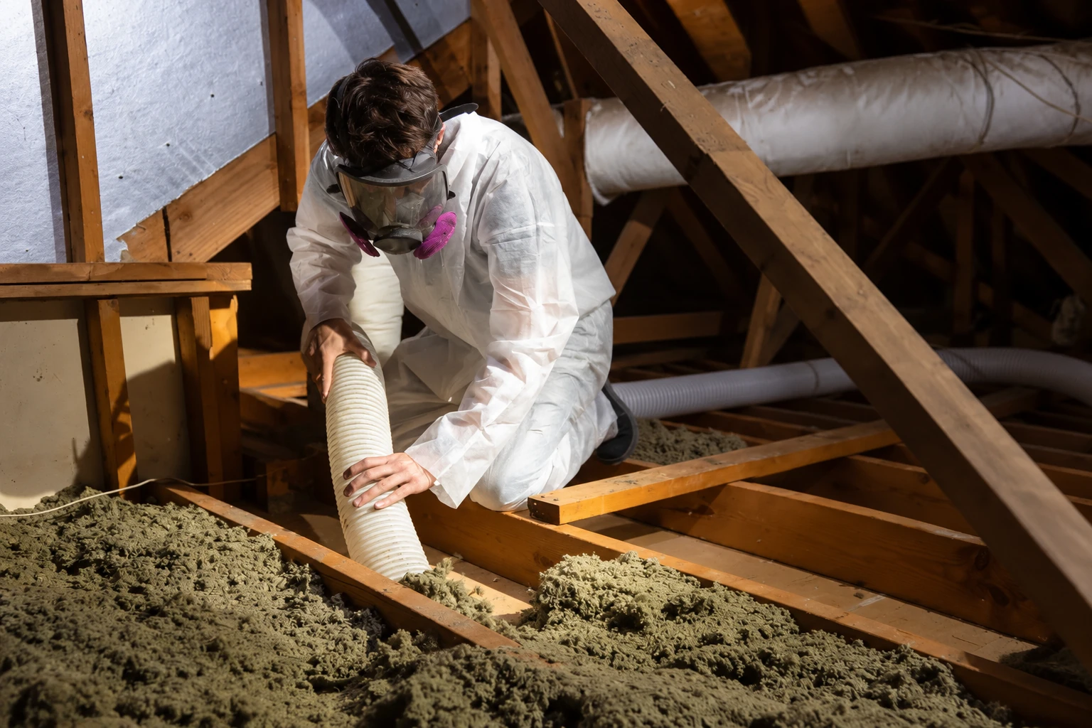 A worker in protective gear uses a vacuum hose to remove loose insulation from an attic.