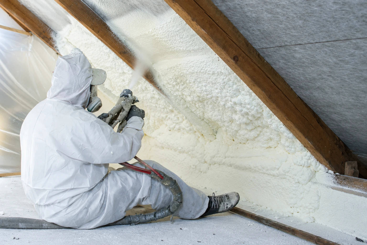 Insulation installer in protective clothing sprays foam onto the underside of a roof between exposed wooden rafters.